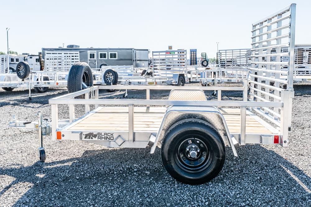 Aluminum utility trailer with wooden deck and single axle, labeled Black Rhino, in a gravel lot among other trailers in a dealership yard.