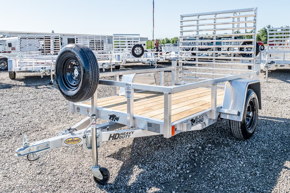Metal utility trailer with a wooden deck and tall slatted loading ramp, parked on a gravel lot. (Informative)