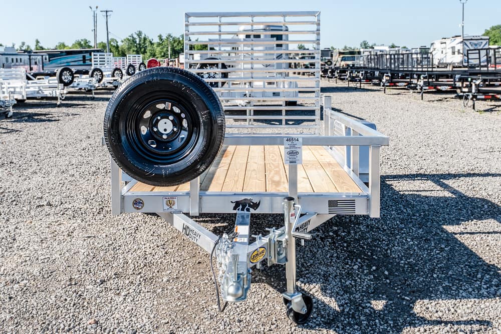 Utility trailer with a wooden deck and a mounted spare tire, parked on a gravel lot among other trailers.