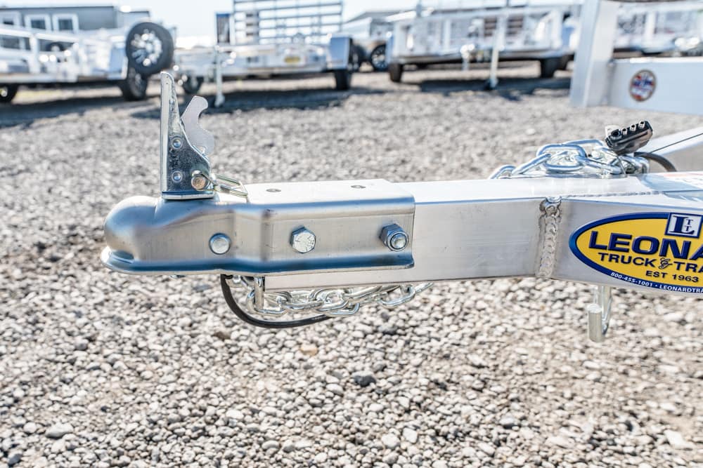 Close-up of a boat trailer hitch with latch, bolts, and safety chains on a gravel lot, with a Leonard trailer sticker nearby.
