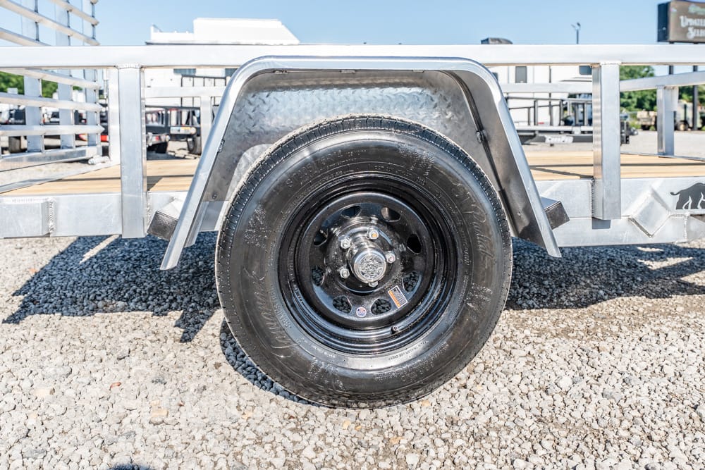 Close-up of a black trailer wheel and tire with a metal fender on a gravel lot.