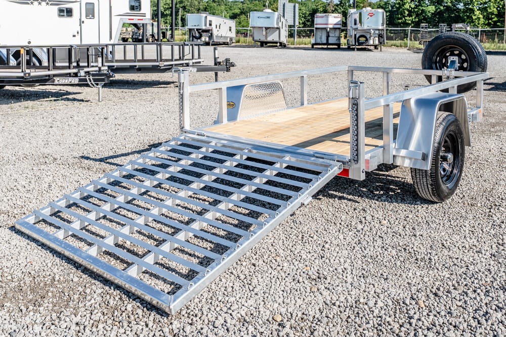 Utility trailer with a lowered metal ramp, wood deck, and single axle, parked on a gravel lot with other trailers in the background.