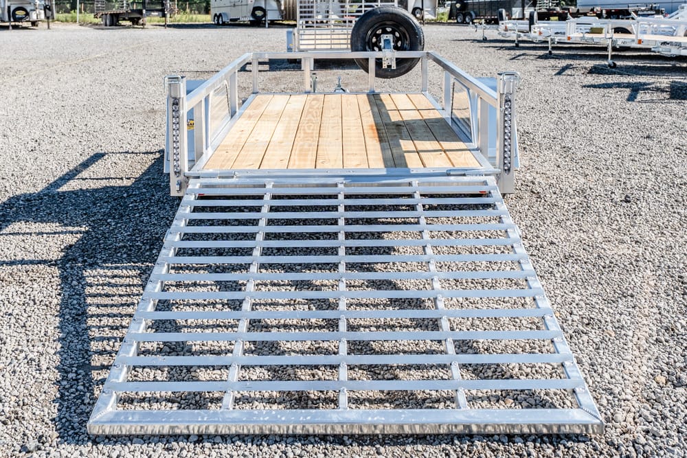 Aluminum utility trailer with a wooden loading deck and a lowered metal ramp on a gravel lot.