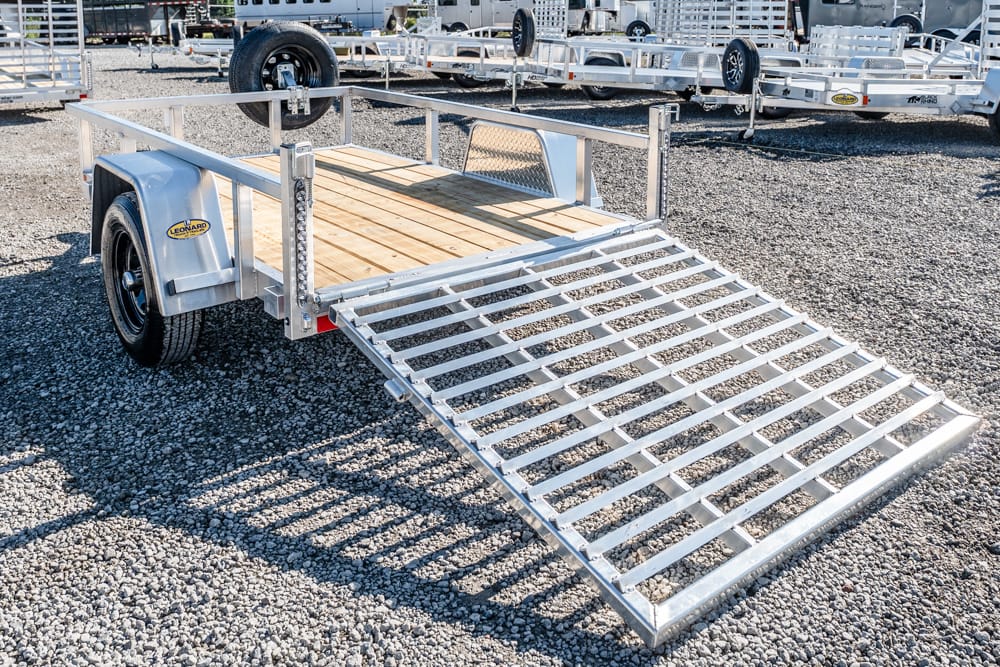 Utility trailer with wooden deck and metal loading ramp lowered, parked on a gravel lot with other trailers nearby, ready for loading or transport.