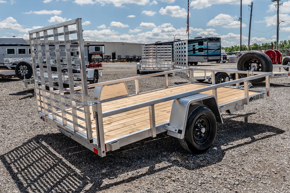 Aluminum flatbed trailer with wooden deck and tall ramp gate on a gravel lot.