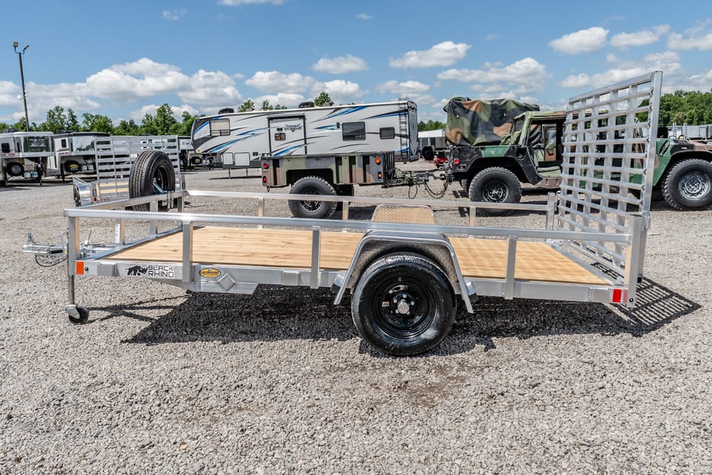 Metal utility trailer with a wooden deck and side rails on a gravel lot, spare tire mounted near the hitch, background shows RVs and campers.