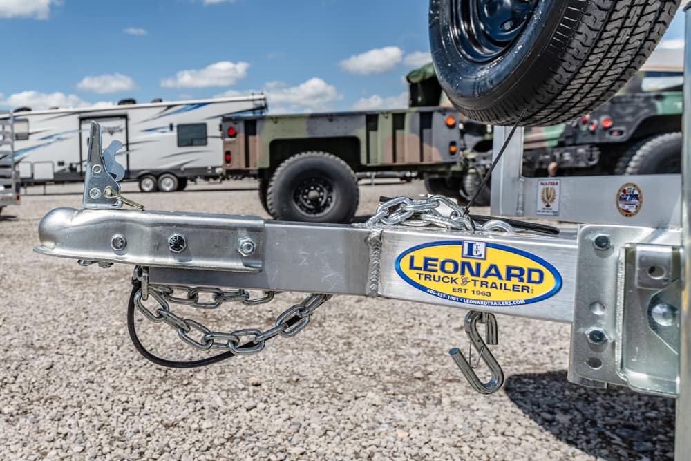Close-up of a aluminum trailer hitch with safety chains, a spare tire above, and a Leonard Truck & Trailer sticker visible on the beam in a gravel lot.