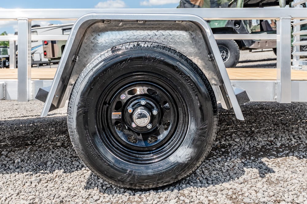 Close-up of a black steel trailer wheel and tire mounted on a silver fender, with gravel ground visible in the foreground.