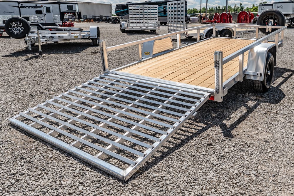Aluminum utility trailer with a fold-down ramp and wooden deck parked on a gravel lot, ready for loading gear or equipment.