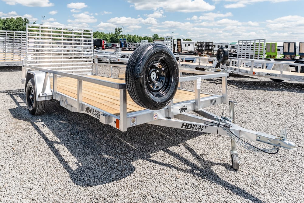 Flatbed utility trailer with a wooden deck and a mounted spare tire, parked on a gravel lot with other trailers in the background.
