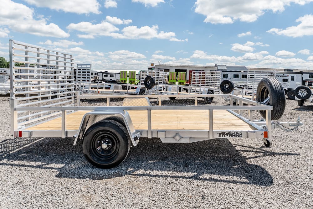 Aluminum-framed flatbed utility trailer with a wooden deck and rear gate, parked on a gravel lot with other trailers in the background, under a partly cloudy sky.