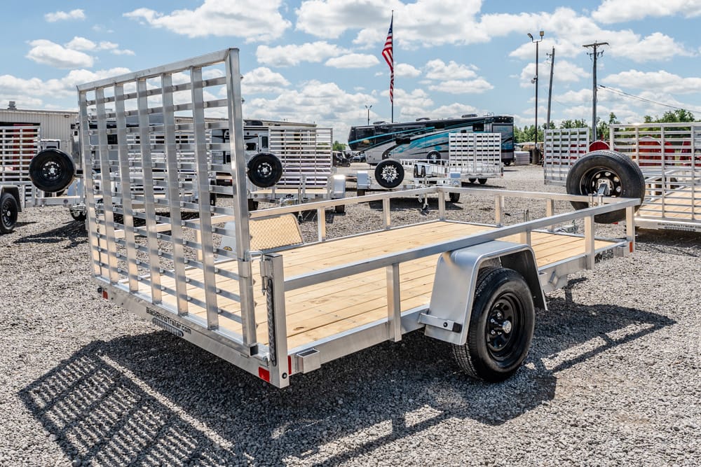 Metal utility trailer with a wooden deck and hinged ramp gate on a gravel lot, with RVs and an American flag in the background.