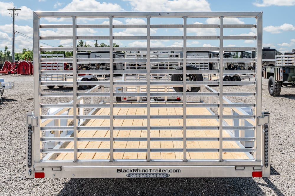 Rear view of an empty aluminum utility trailer with a wooden deck and horizontal bars, parked in a gravel lot at a trailer dealership (BlackRhinoTrailer.com)
