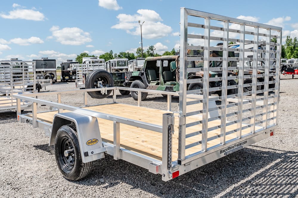 Aluminum flatbed utility trailer with a wooden deck in a gravel lot, back ramp gate raised in a trailer yard.