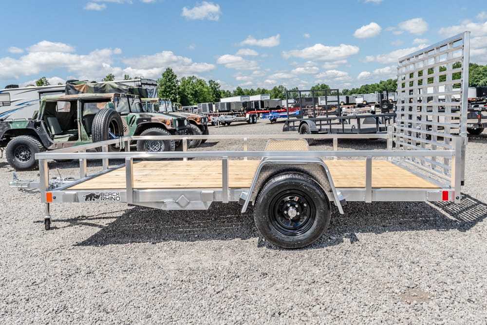 Flatbed utility trailer with a wooden deck and side rails, parked on a gravel lot (Black Rhino logo visible).