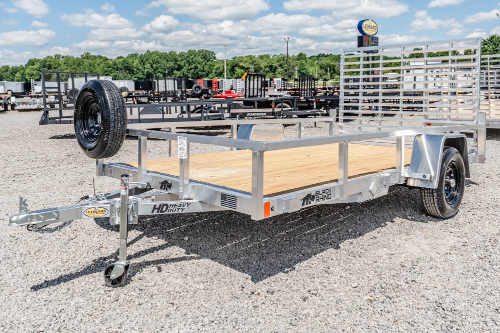 Flatbed utility trailer with a wooden deck, spare tire mounted on the front left, and a tall metal ramp gate at the rear; single-axle fenders with a gray metal frame and branding visible (HD Heavy Duty, Black Rhino).