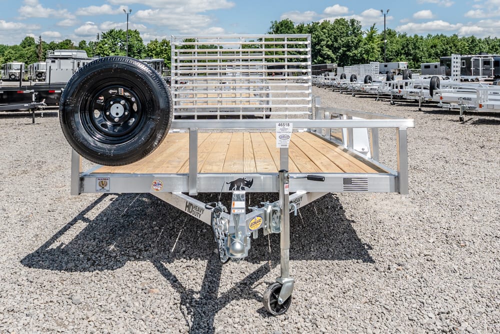 Flatbed utility trailer with a wooden deck and a spare tire on the left, parked in a gravel trailer lot.