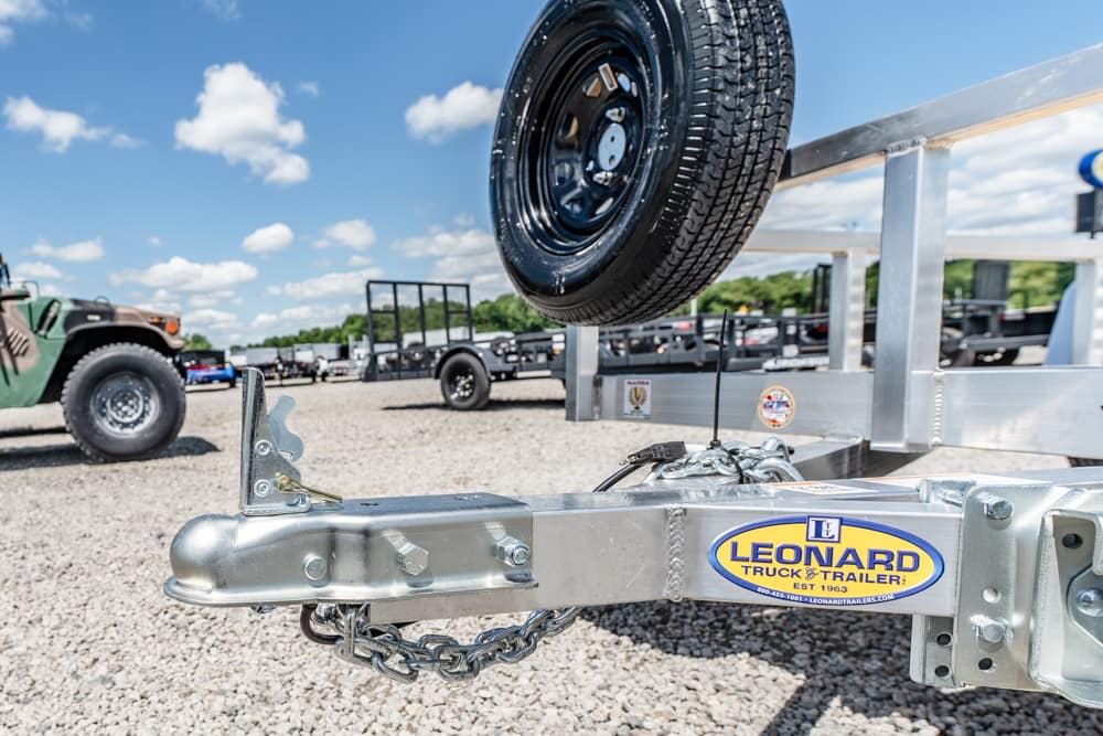 Close-up of a galvanized trailer hitch with safety chains, spare wheel above, on a gravel lot at a Leonard Truck & Trailer lot.