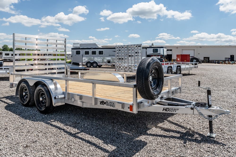 Aluminum flatbed utility trailer with a wooden deck and spare tire mounted at the front, on a gravel lot with RVs in the background.
