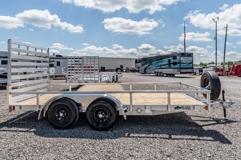 Silver dual-axle flatbed trailer on a gravel lot with wooden deck and metal rails, ready for hauling and with a spare tire at the hitch.