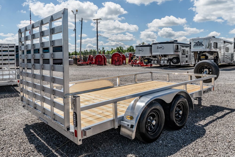 Aluminum flatbed trailer with a wooden deck and raised rear gate on a gravel lot, ready for loading.
