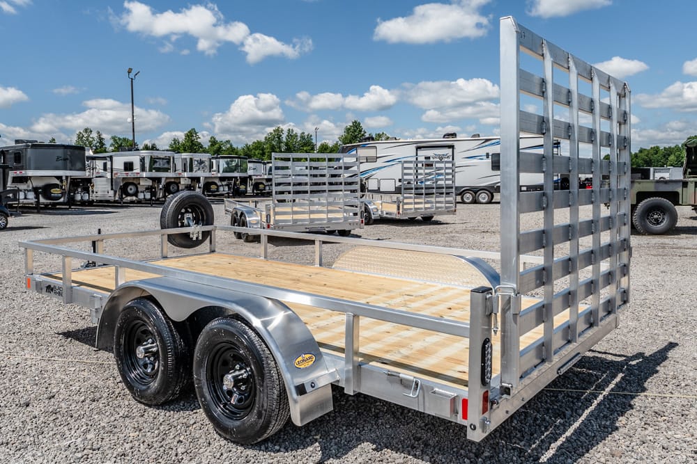 Dual-axle aluminum utility trailer with a wooden deck and a tall rear ramp gate in a gravel trailer lot.
