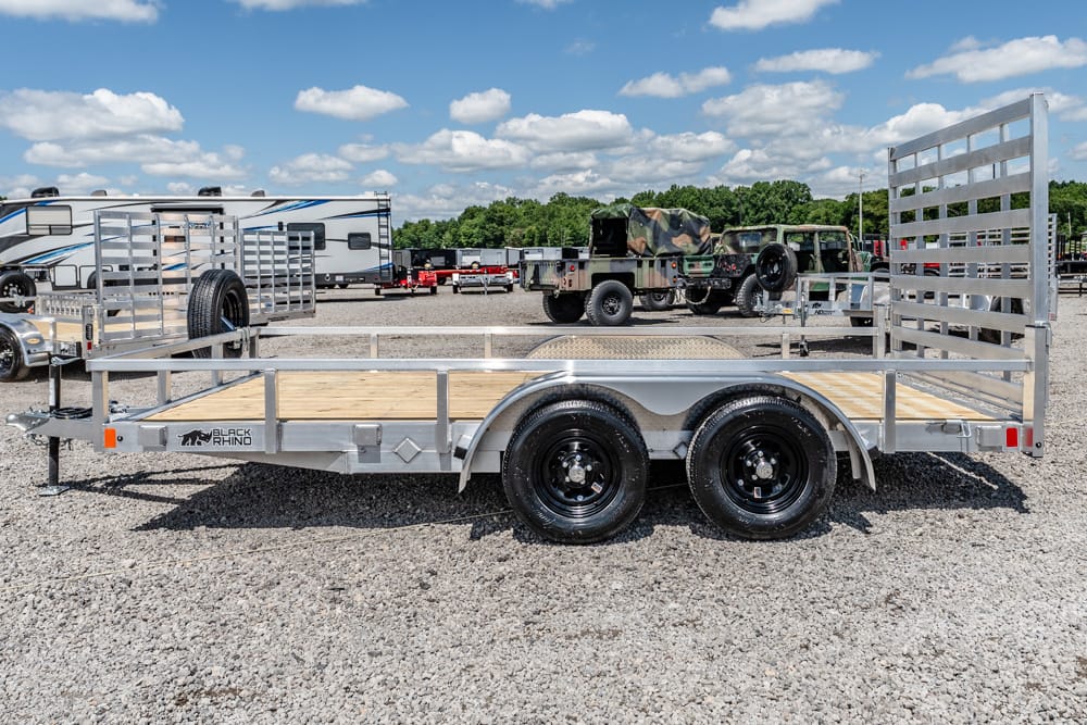 Dual-axle utility trailer with wooden deck, spare tire at the front, and rear ramp gate on a gravel lot.