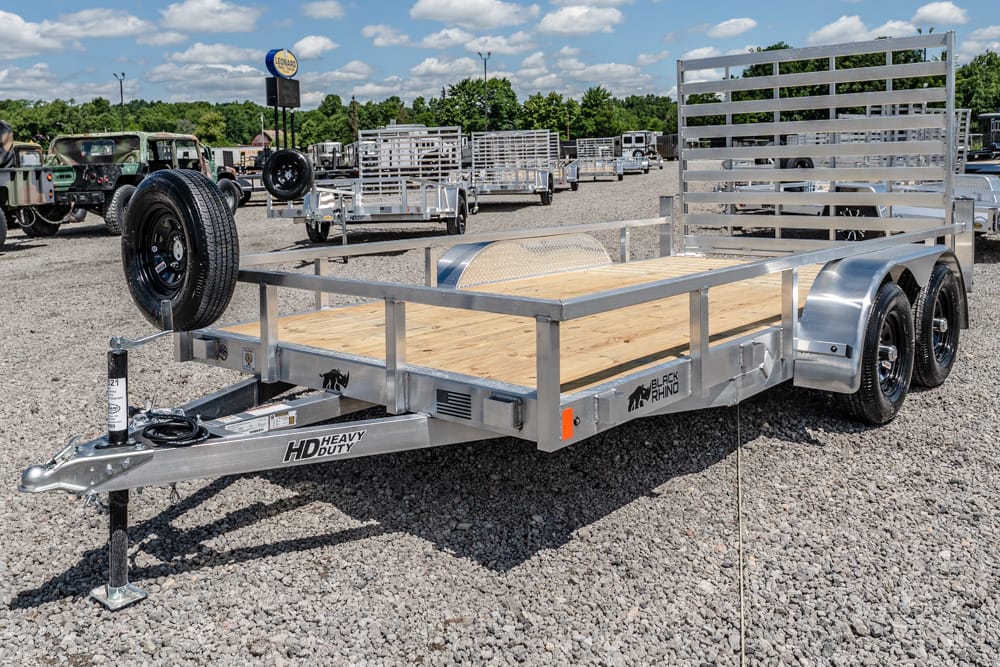 Aluminum utility trailer with a wooden deck, dual axles, a spare tire on the front left, and a tall slatted rear ramp in a gravel trailer lot.