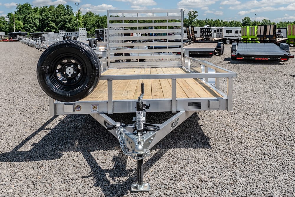 Flatbed utility trailer with wooden deck and spare tire mounted on the side in a gravel lot.