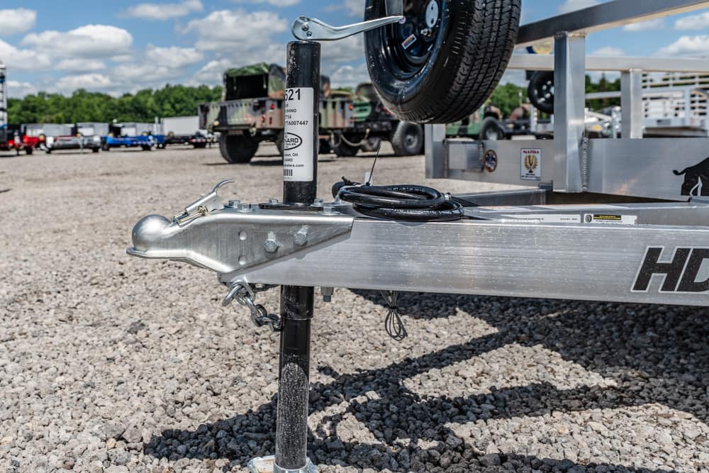 Close-up of a boat trailer hitch and jack on a gravel lot, with a spare tire mounted on the trailer in the background.