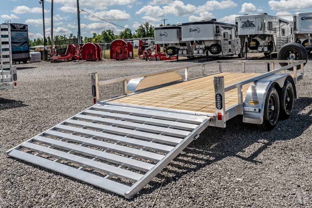 Aluminum flatbed utility trailer with a lowered ramp at ground level in a gravel lot, RVs and trailers in the background.