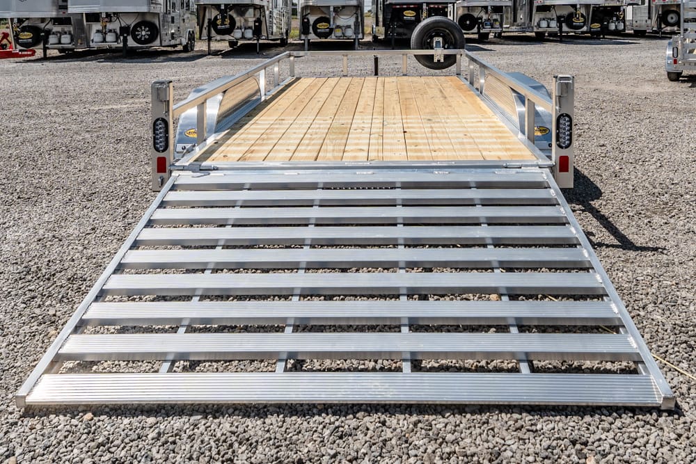 Metal trailer ramp with slatted aluminum sides and wooden deck, extended on a gravel lot for loading near a row of trucks