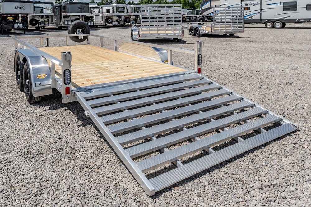 Silver utility trailer with a wooden deck and lowered metal ramp on a gravel lot, surrounded by other trailers and RVs