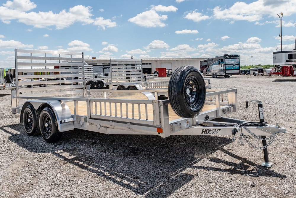 Aluminum dual-axle utility trailer with wooden deck and a mounted spare tire, parked on a gravel lot with RVs in the background.