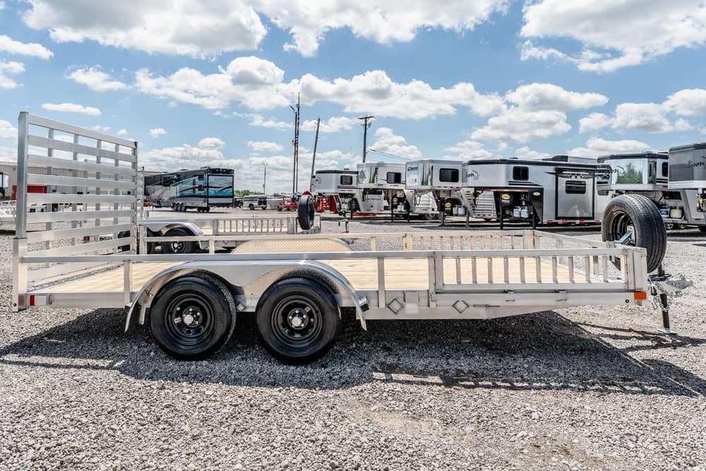 Aluminum double-axle utility trailer with a wooden deck, parked on a gravel lot with RV trailers in the background.