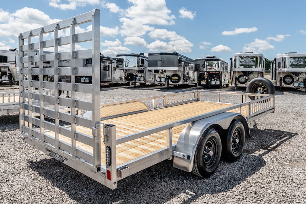 Silver dual-axle utility trailer with a wooden deck and tall tailgate, parked on a gravel lot with horse trailers in the background