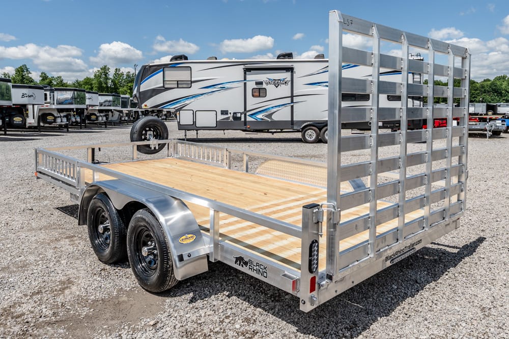 Dual-axle utility trailer with a wooden bed and metal side rails parked on gravel, with a large RV in the background.