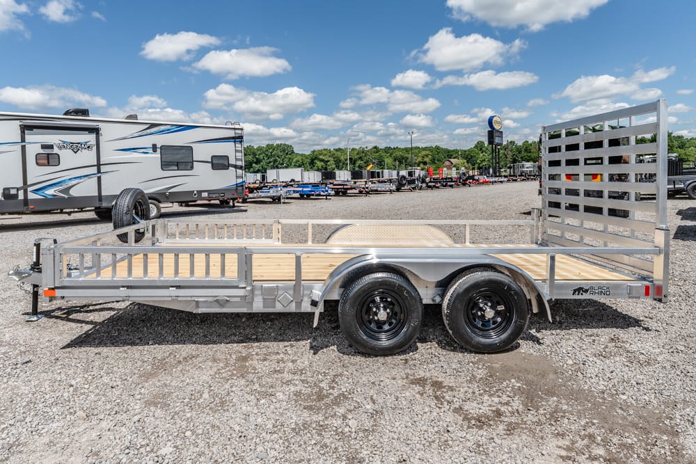 Twin-axle utility trailer with a wooden deck and metal rails, parked on a gravel lot with an RV in the background