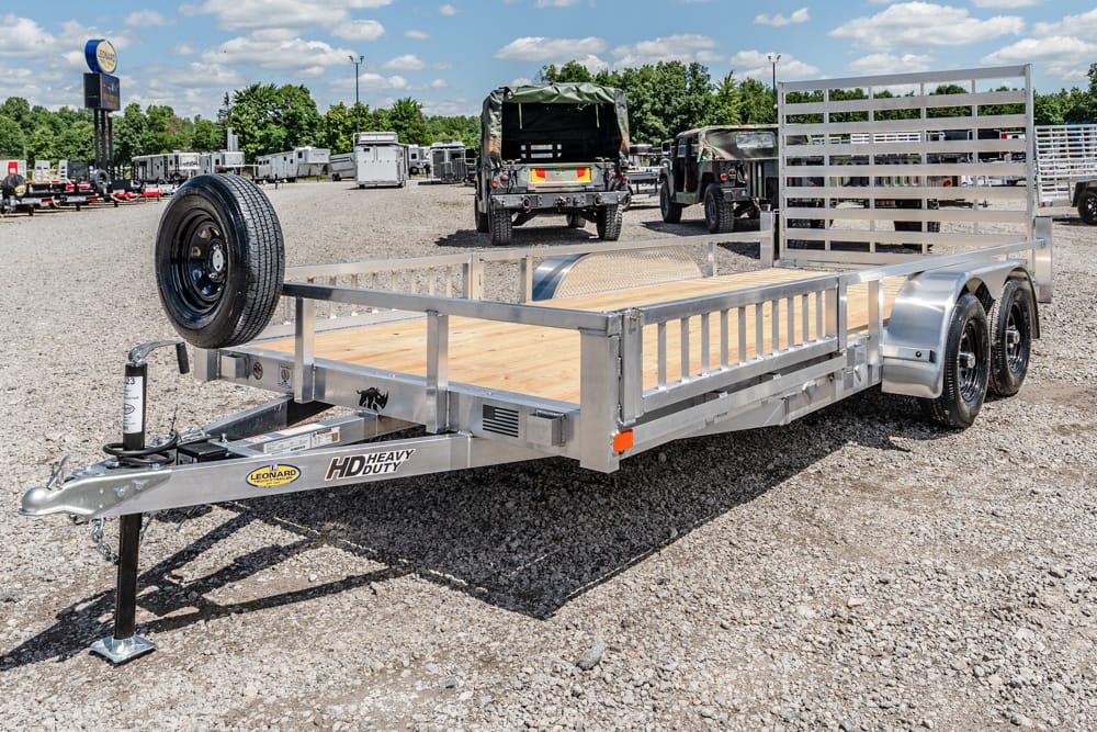Dual-axle utility trailer with a wooden deck and spare tire, parked on a gravel lot with other trailers nearby.