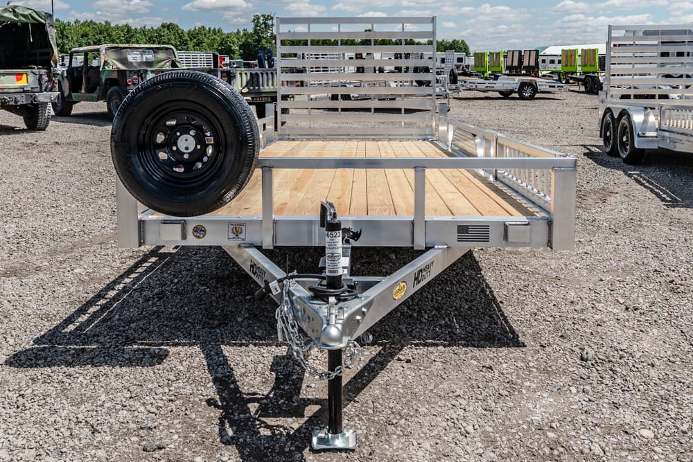Flatbed utility trailer with a wooden deck and a spare tire mounted on the left, parked on a gravel lot with other trailers in the background.