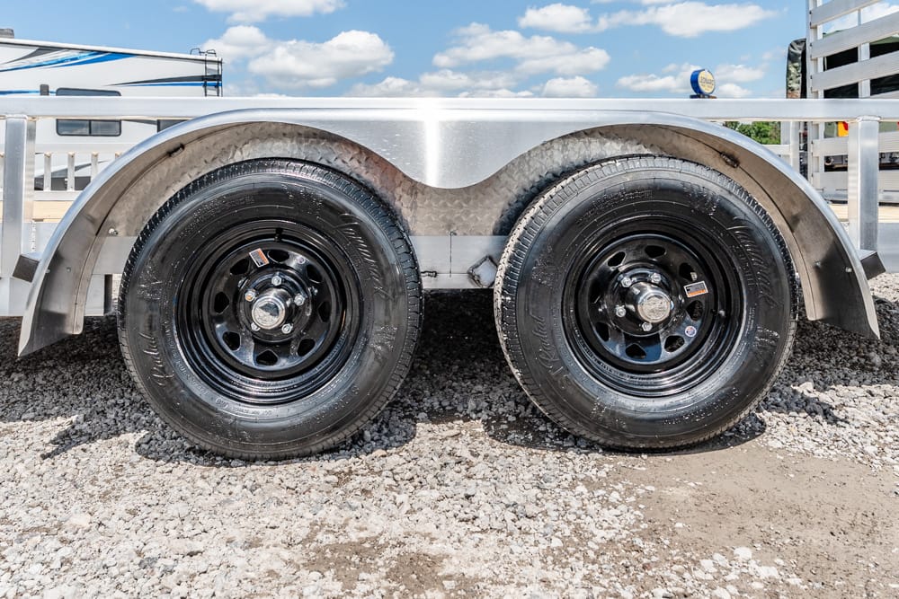Dual trailer wheels with black steel rims under a curved metal fender on a gravel lot in daylight.