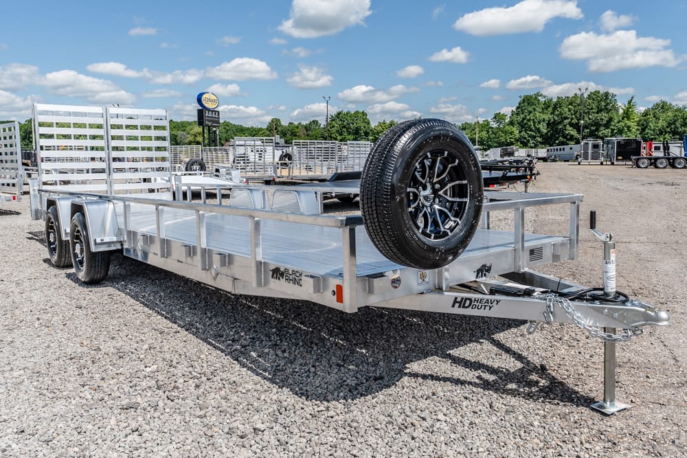 Aluminum heavy-duty dual-axle trailer with a spare tire mounted on the front deck in a gravel lot