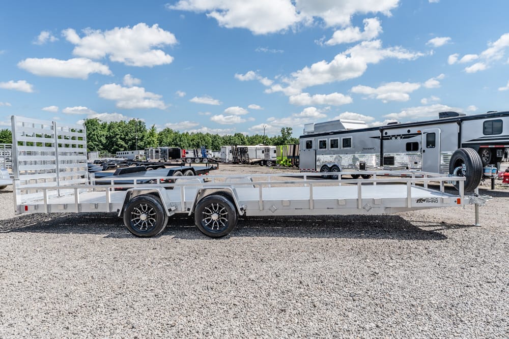 Aluminum utility trailer with dual axles and a tall rear ramp, parked in a gravel lot with RVs in the background