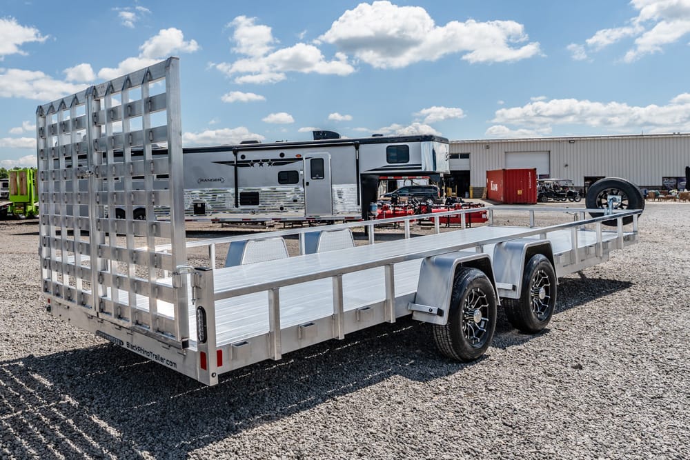 Aluminum dual-axle utility trailer with a tall ramp at the back, parked on a gravel lot with RVs in the background.