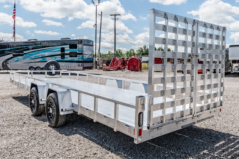 Aluminum flatbed trailer with a tall rear ramp gate on a gravel lot, with a large RV in the background.