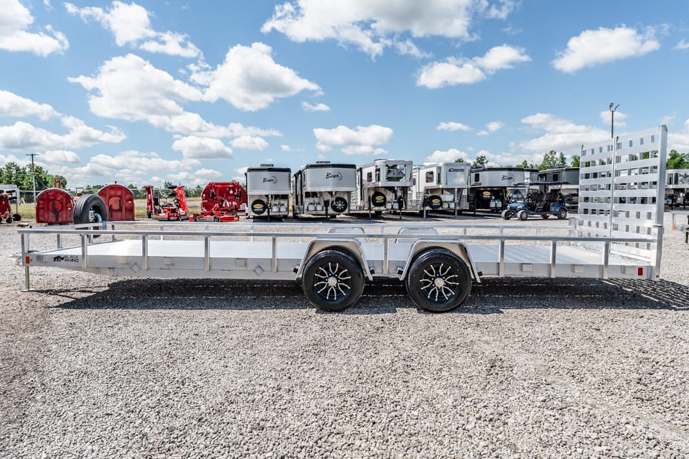 Aluminum flatbed trailer with dual axles parked on a gravel lot, with travel trailers in the background under a blue sky.