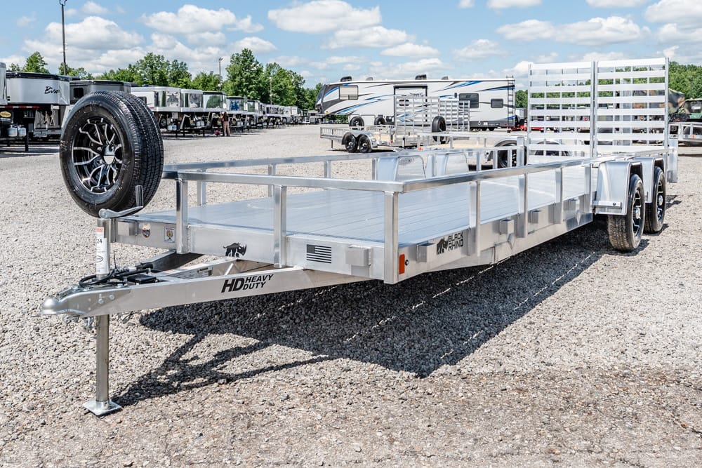 Open, aluminum flatbed trailer with dual axles and a mounted spare tire at the front, parked on a gravel lot among other trailers and RVs.",