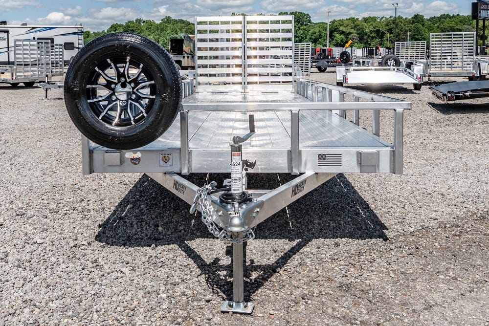 Front view of an aluminum utility trailer with a spare tire mounted on the left side and a hitch in a gravel lot.