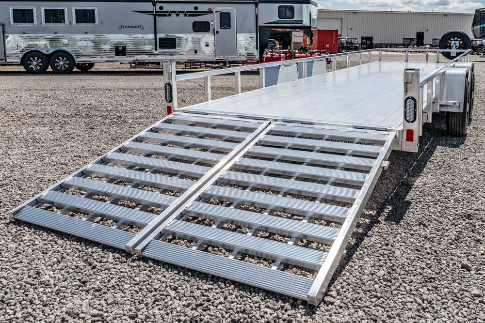 Aluminum flatbed trailer with two folding loading ramps extended on a gravel lot, with another trailer in the background.