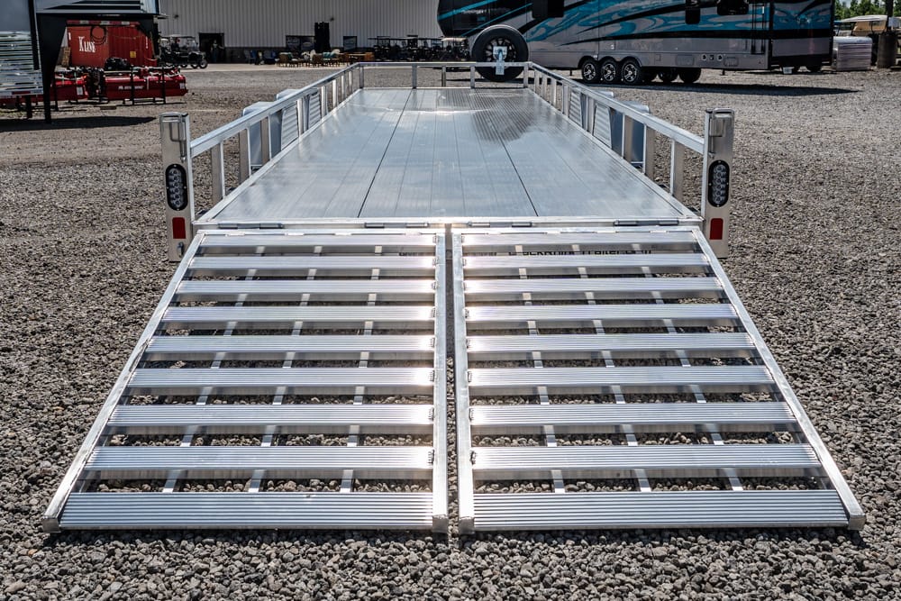Aluminum flatbed trailer with ramp gates folded down on a gravel lot, with RVs in the background.
